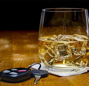 A glass of whiskey with a car key next to it on a wooden table - John Abel Law Office